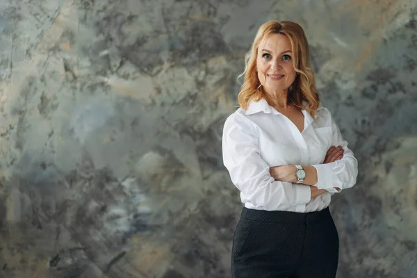 Ofisteki beyaz tişörtlü güzel olgun bir kadın koyu renk arka planda duruyor ve kollarını kavuşturup kameraya bakıyor. Başarılı bir olgun kadın.