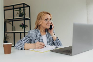 Güzel yaşlı iş kadını çalışırken ve not defterine notlar yazarken akıllı telefonuyla konuşuyor. Olgun kadın 50 'li yaşlarda.