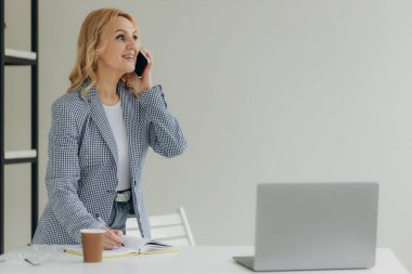 Güzel yaşlı iş kadını çalışırken ve not defterine notlar yazarken akıllı telefonuyla konuşuyor. Olgun kadın 50 'li yaşlarda.