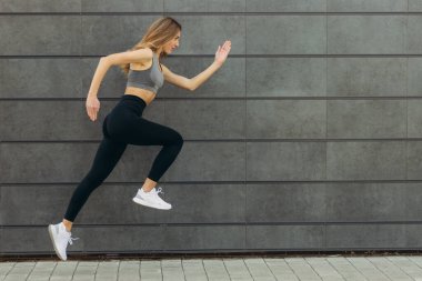 Zıplayan ve koşan zinde, genç, sportif, güzel bir kadın, gri duvar arkasına doğru acele edin. Jimnastikçi, dişlek gülümsemesiyle, uzun boyuyla, açık havada yüksekten atlıyor.