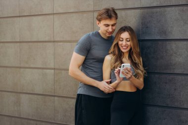 Gülümseyen sportif çift gri duvarda cep telefonu kullanıyor..