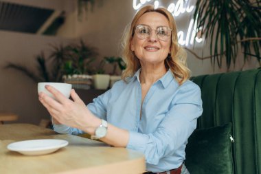 Kafede kahve içen güzel, gülümseyen bir kadın. Kafeteryada sıcak kapuçino içen olgun bir kadının portresi şık kıyafetler giyer.