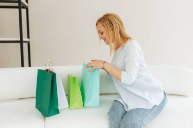 Kanepede oturan olgun bir kadın marketten gelen paketlerden birine bakıyor. Odada bir sürü renkli çanta var. Çevrimiçi alışveriş kavramı