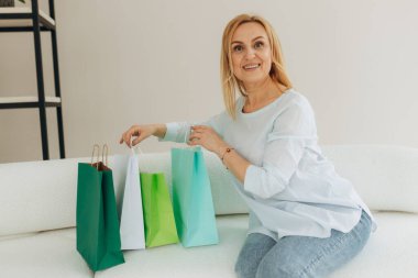 Kanepede oturan olgun bir kadın marketten gelen paketlerden birine bakıyor. Odada bir sürü renkli çanta var. Çevrimiçi alışveriş kavramı