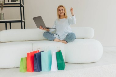 Neşeli, güzel, beyaz, olgun, sarışın bir kadın, yeni alınmış kıyafetlerle mutlu, evdeki bir koltukta oturuyor, online ödemeler için laptop ve kredi kartını kullanıyor, yanında bir sürü kağıt torba, gülümsüyor.