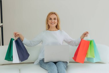 Portre mutlu olgun kadın alışveriş çantalarını evde tutuyor. İnternetten alışveriş. İnternetten laptop alışverişi yapan kıdemli bir kadın.