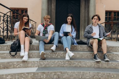 Study together is fun, teamwork, team building concept. Four happy students are sitting near college building and holding books, devices, wearing casual smart, smiling, on a nice summer