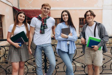 Study together is fun, teamwork, team building concept. Four happy students are sitting near college building and holding books, devices, wearing casual smart, smiling, on a nice summer