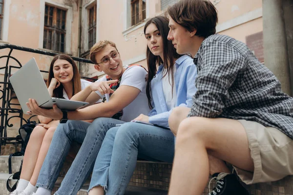 Study together is fun, teamwork, team building concept. Four happy students are sitting near college building and holding books, devices, wearing casual smart, smiling, on a nice summer