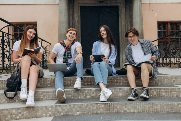 Study together is fun, teamwork, team building concept. Four happy students are sitting near college building and holding books, devices, wearing casual smart, smiling, on a nice summer