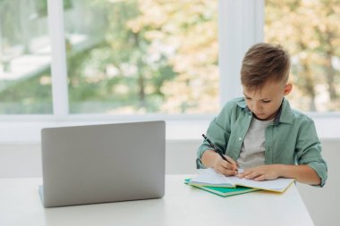 Adorable attentive boy writing in notebook and using laptop while doing homework. Place for text