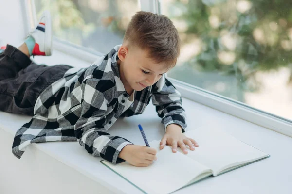 Çocuk ev ödevi yapıyor. Çocuk pencere eşiğine uzanıyor ve bir deftere yazıyor. Öğrenci çocuk pencere kenarında ödev yapıyor. Evde eğitim.