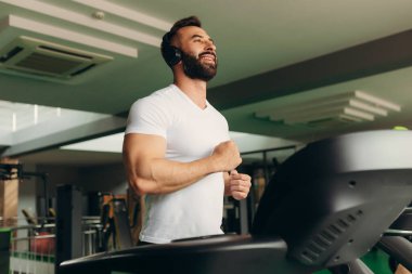 Side view of a handsome athlete running on a treadmill in the gym. The guy is wearing a white shirt