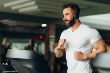 Side view of a handsome athlete running on a treadmill in the gym. The guy is wearing a white shirt