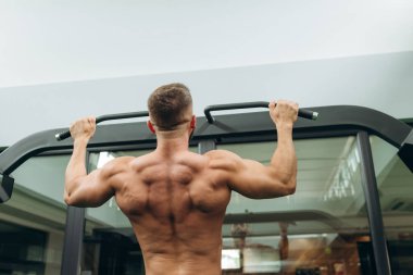 Handsome fitness man doing back workout using horizontal bar at gym, rear view. Young Athlete Doing Pull Ups