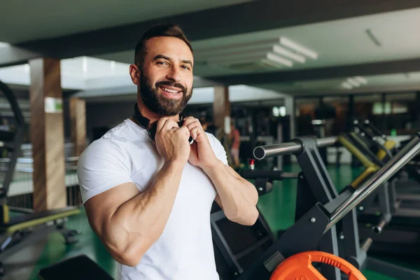 Smiling young man in white t-shirt puts on headphones to listen to music in the gym while doing exercises and lifting weights