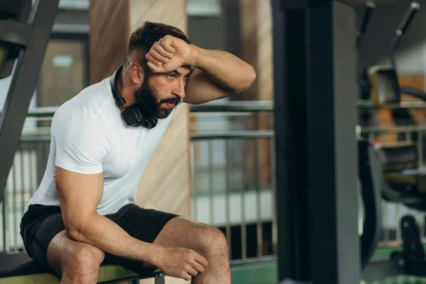 A young man wipes sweat from his forehead in the gym. He is tired and exhausted from weight lifting and strength training.