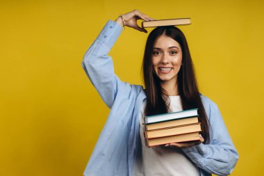 Portrait of student girl balancing books on head and hand while standing against yellow background