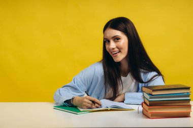 Beautiful smiling woman sitting at table, writing in her notebook on yellow background. Education concept.