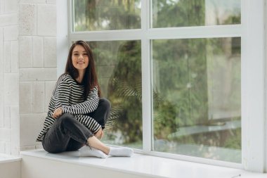 Smiling woman in sitting on windowsill looking at camera