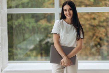 A young woman stands near a window with a view of nature and holds a closed laptop in her hands