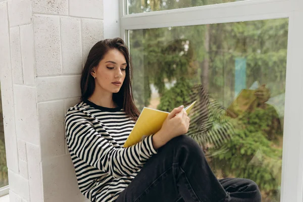 Young caucasian girl sits on windowsill in bright room and enthusiastically reads book in good lighting. Student with dark hair is studying literature during summer holidays. Free time concept