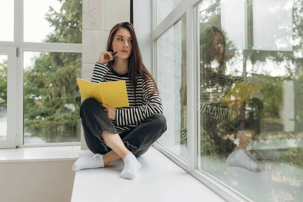 Home schooling. A teenager girl, a student, sits on a windowsill writes homework. Distance learning, preparation for exams. Soft selective focus.