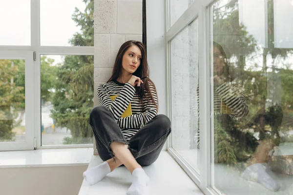 A beautiful happy young woman is sitting on the windowsill with a book in her hands