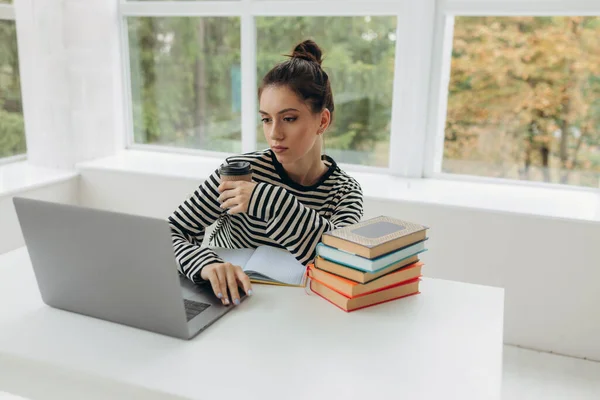 Portrait of young beautiful smiling young woman sitting at office desk enjoying her cup of coffee while working or studying on laptop computer at small home office or in the student dorm. Indoors