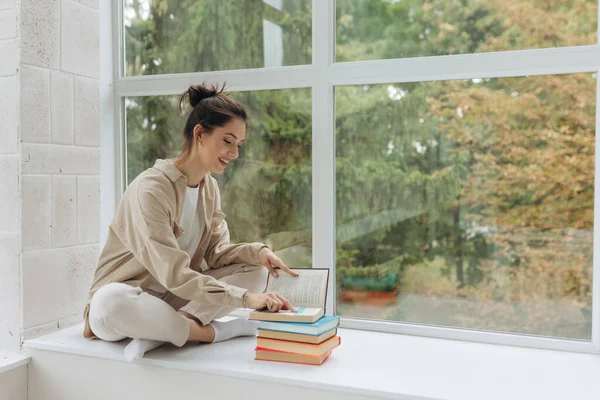 Young beautiful girl sitting on windowsill, holding and reading book