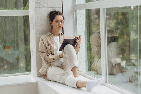 Beautiful young woman sitting by the window and reading a book.