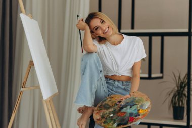 Positive young woman holding paintbrush and palette with colorful paints near easel. Artist