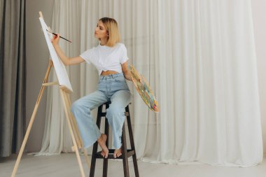 Positive young woman holding paintbrush and palette with colorful paints near easel. Artist