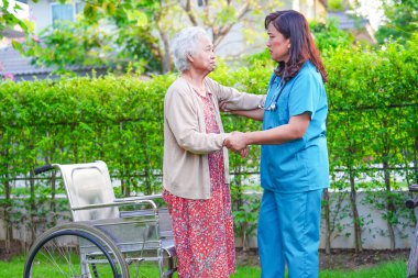 Bakıcı, parkta tekerlekli sandalyede oturan Asyalı yaşlı kadın maluliyet hastasına yardım ediyor..