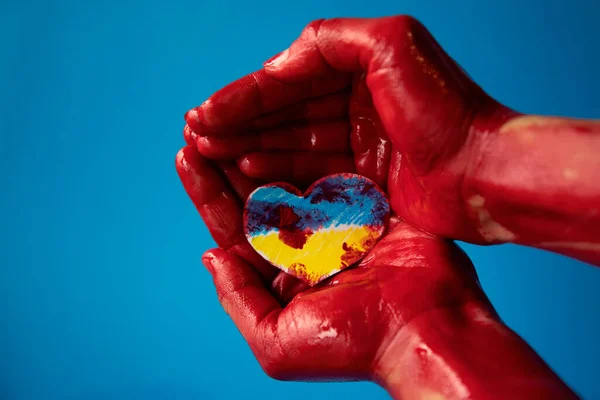 Ukrayna bayrağı renginde ve kanlı savaş konseptiyle boyanmış bir kalbin kağıt kesiğini tutan mavi arka planda bir kadının elleri..