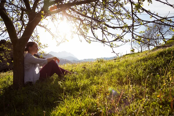 Genç esmer kız gün batımında bir ağacın gövdesine yaslanmış sonsuzluğa bakıyor. Kırsal alan, arka planda dağlar olan çimenlik alan. Özgür ve vahşi. Rahatla konsept