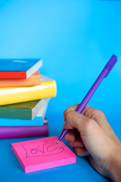 Yığılmış kitapların dikey fotoğrafı lgtb bayrağının renkleri ve bir el postit üzerine aşk yazıyor. kopyalama alanı ve mavi arkaplan. Ayrımcılık yok,