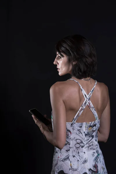 portrait of young woman with black hair absentmindedly looking at the mobile phone she is holding on a black background