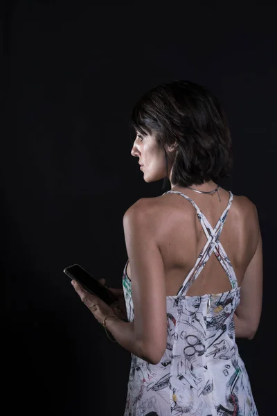 portrait of young woman with black hair absentmindedly looking at the mobile phone she is holding on a black background