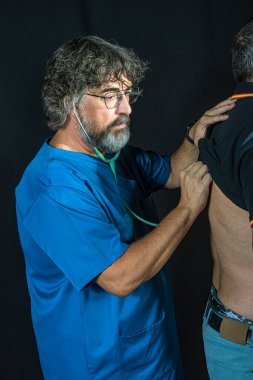 Sakallı, mavi ameliyat odası elbisesi giymiş, siyah bir geçmişi olan olgun bir doktor hasta sağlığı mesleğini inceliyor.