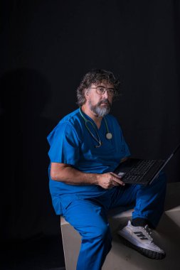 Mavi ameliyathane kıyafeti giymiş sakallı olgun bir doktor ahşap bir kutunun üzerinde oturmuş bir bilgisayar sağlığı uzmanına danışıyor.