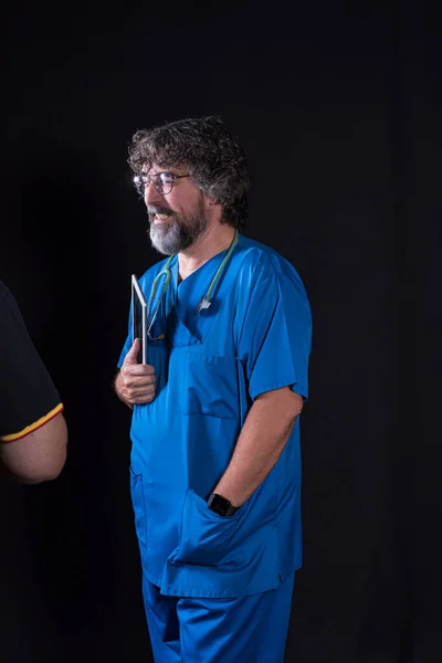 mature doctor with a beard dressed in a blue operating room suit on a black background holding a tablet looking at the camera healthcare medical professions