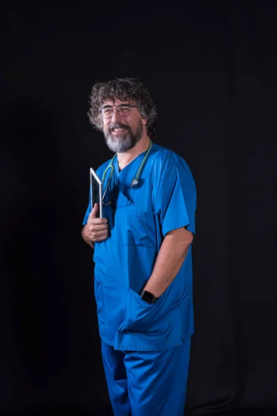 mature doctor with a beard dressed in a blue operating room suit on a black background holding a tablet looking at the camera healthcare medical professions