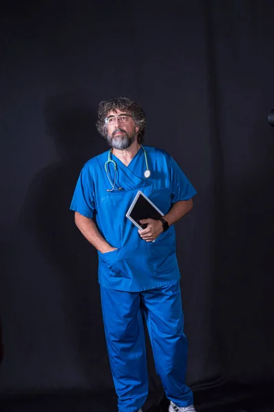 mature doctor with a beard dressed in a blue operating room suit on a black background holding a tablet looking at the camera healthcare medical professions