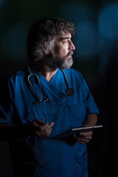 mature doctor with a beard dressed in a blue operating room suit on a black background consulting a tablet healthcare medical professions