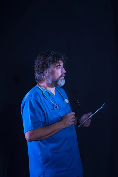 mature doctor with a beard dressed in a blue operating room suit on a black background consulting a tablet healthcare medical professions