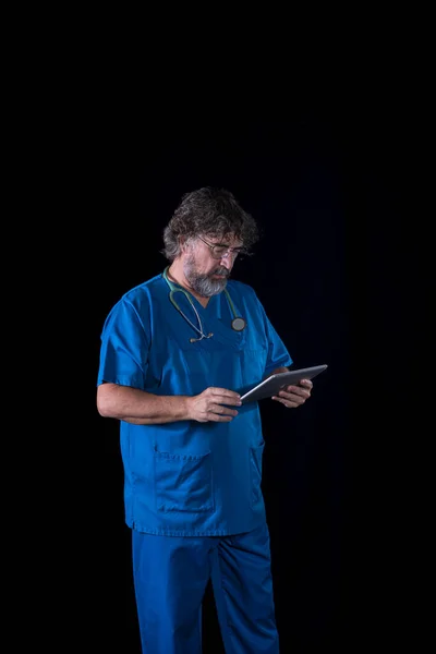 mature doctor with a beard dressed in a blue operating room suit on a black background consulting a tablet healthcare medical professions