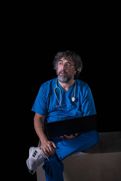 bearded mature doctor dressed in blue operating room suit sitting on a wooden box consulting a computer healthcare medical professions