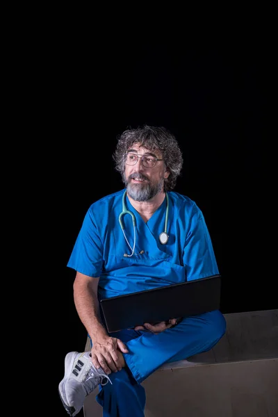 bearded mature doctor dressed in blue operating room suit sitting on a wooden box consulting a computer healthcare medical professions