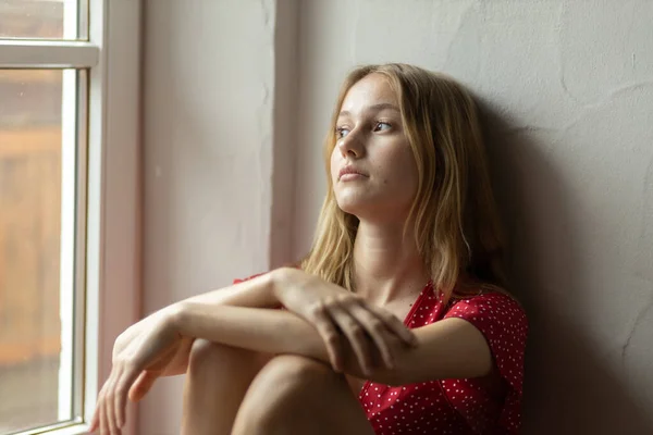 A romantic girl in a red dress sits on the floor near the window and looks out the window. High quality photo